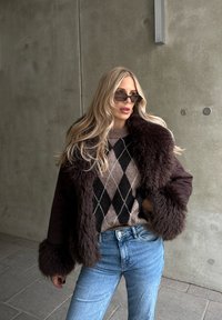 Hnedý kabát z umelej kožušiny so širokými manžetami, nosený cez béžový a čierny argyle sveter; kombinovaný so svetlomodrými džínsami a tmavými slnečnými okuliarmi.