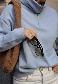 Pull bleu clair à fermeture éclair avec col entonnoir et poignets côtelés, associé à un sac brun texturé. Tenant des lunettes de soleil foncées aux verres verts.