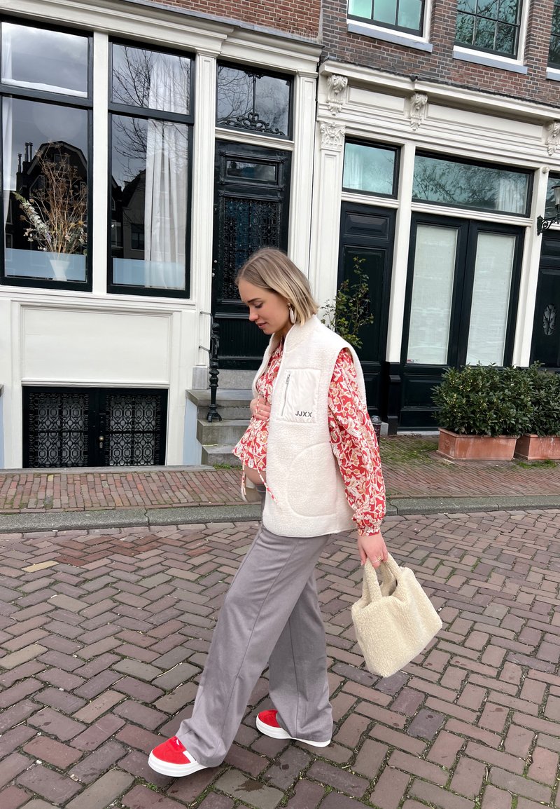 Λευκό γούνινο γιλέκο, κόκκινο φλοράλ μπλουζάκι με μακριά μανίκια, γκρι παντελόνι φαρδύ, κόκκινα αθλητικά παπούτσια, κρατώντας μια υφασμάτινη μπεζ τσάντα, περπατώντας σε πλακόστρωτο.