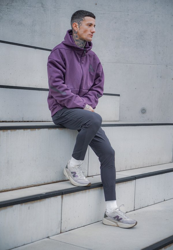 Purple zip-up hoodie with a large front pocket, paired with slim-fit dark trousers and white socks. Features light grey sneakers with purple accents.