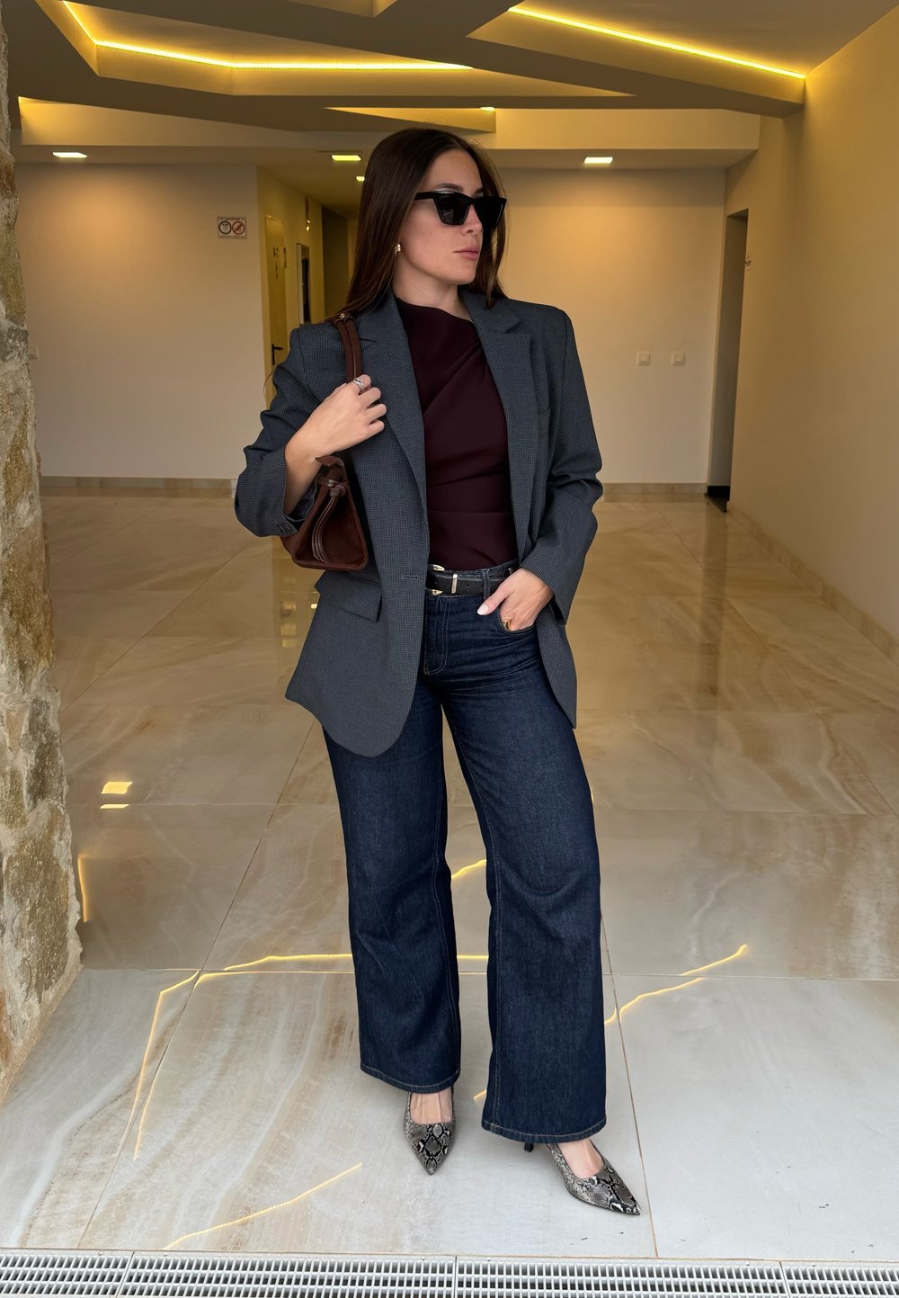 Blazer à carreaux gris, haut bordeaux, jean flare en denim foncé et talons à imprimé serpent. Sac à main marron et lunettes de soleil complètent le look.