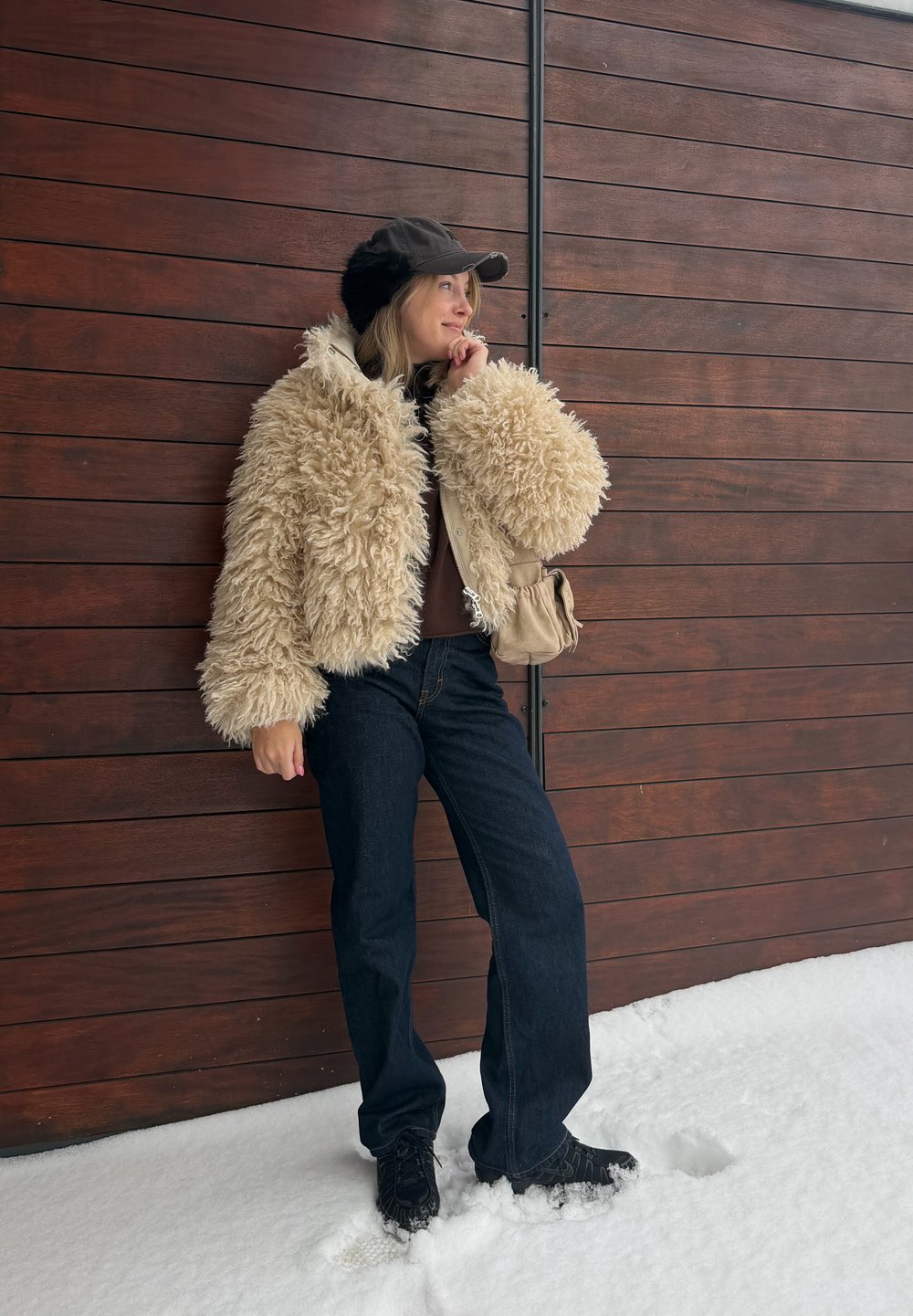 Femme en veste beige duveteuse, casquette noire avec cache-oreilles, jean foncé et chaussures noires, debout sur la neige près d'un mur en bois.