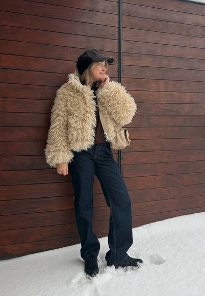 Mujer con chaqueta beige y esponjosa, gorra negra con orejeras, vaqueros oscuros y zapatos negros, de pie sobre la nieve junto a una pared de madera.