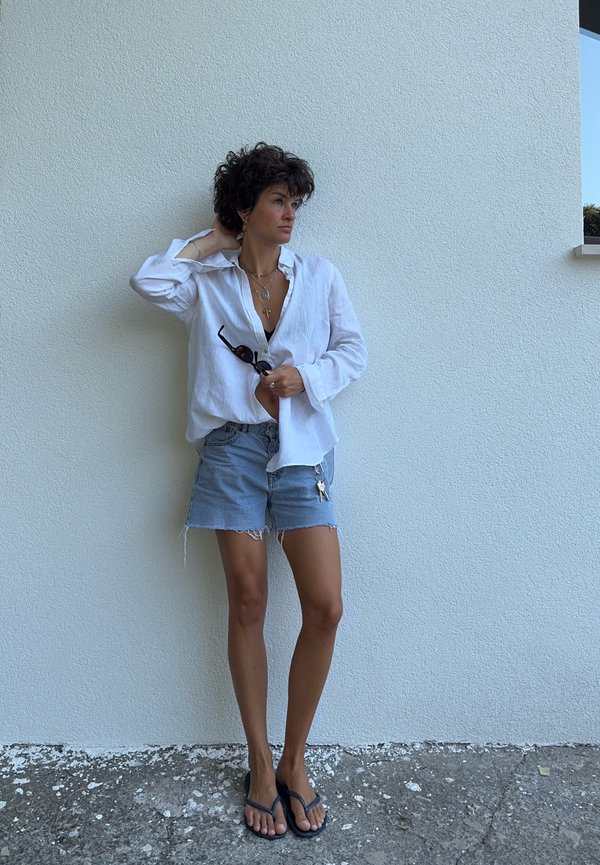 Donna con capelli ricci che indossa una camicia bianca, pantaloncini di jeans e infradito, in piedi contro un muro bianco, tiene gli occhiali da sole e si tocca i capelli.
