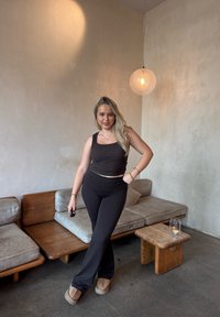 Sort tanktop og sort bukser med høj talje på en person, der står ved siden af træmøbler og en glaslysholder i et minimalistisk rum.