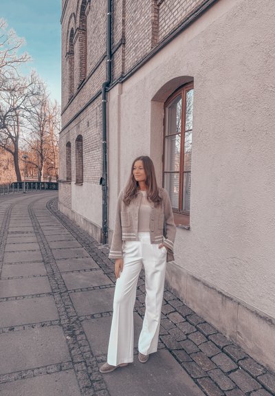 Chaqueta de corte corto gris claro, blusa beige y pantalones anchos blancos. Tejido suave y líneas limpias, de pie junto a un edificio de ladrillo con pavimento.