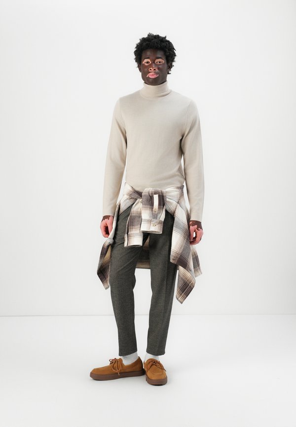 Beige rullekrave sweater, grå plaid flannel skjorte knyttet om taljen, grå bukser og brune snøresko, stående foran en hvid baggrund.