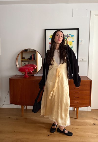 Mujer con vestido amarillo y chaqueta negra sosteniendo una bolsa negra, de pie frente a una cómoda de madera con una lámpara roja y una obra de arte enmarcada y colorida.