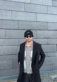 Cappotto nero sopra una camicia a righe con tasche verde menta, cappello nero e occhiali da sole, sullo sfondo di un muro grigio texturizzato.