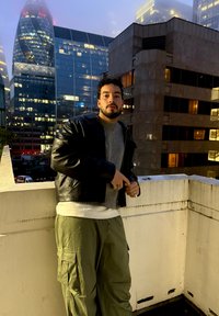 Fekete bőrkabát szürke pulóver felett, laza olívazöld cargo nadrággal párosítva. Városi háttér üveg felhőkarcolókkal és lágy megvilágítással.