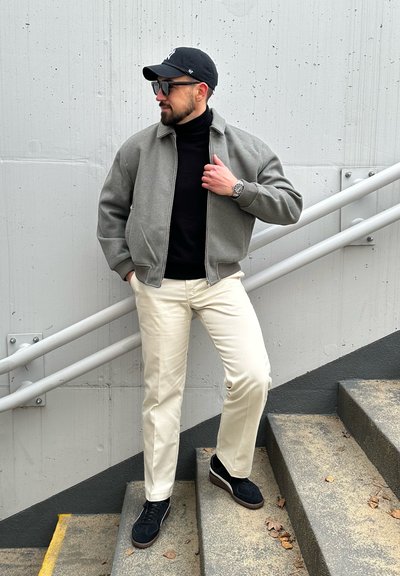 Chaqueta bomber gris sobre un suéter de cuello alto negro, pantalones de color crema y zapatillas negras. De pie en unas escaleras con un fondo de pared de concreto.