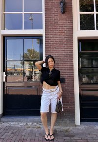 Zwarte cropped mesh top, witte denim shorts met kettingaccent, flip-flops en een kleine bruine tas met patroon. Bakstenen achtergrond met deuren.