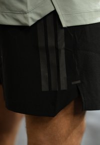 Gros plan sur une personne portant un short de sport noir avec trois rayures verticales foncées sur le côté et une petite fente latérale.