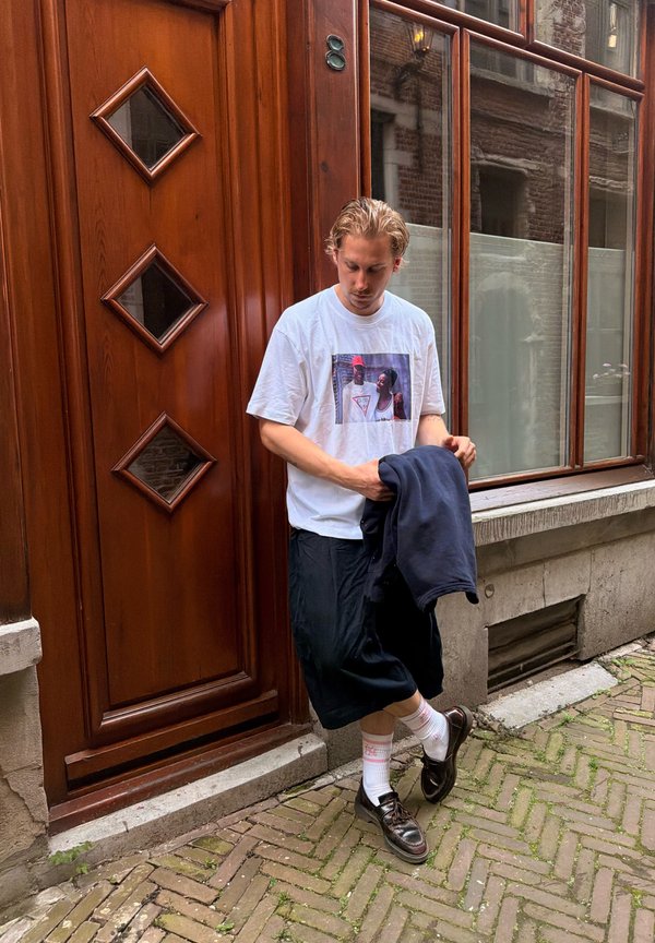 Jonge man met licht haar, gekleed in een wit T-shirt met opdruk, zwarte korte broek, witte sokken en zwarte schoenen, staat bij een houten deur en raam op een kinderkopjesstraat.