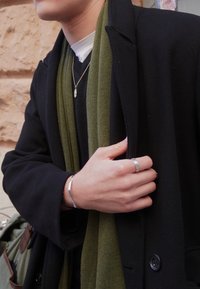 Άτομο με μαύρο παλτό, πράσινο κασκόλ, ασημένιο κολιέ, δαχτυλίδι και βραχιόλι, κρατώντας το παλτό κοντά στο στήθος, σε εξωτερικό χώρο.