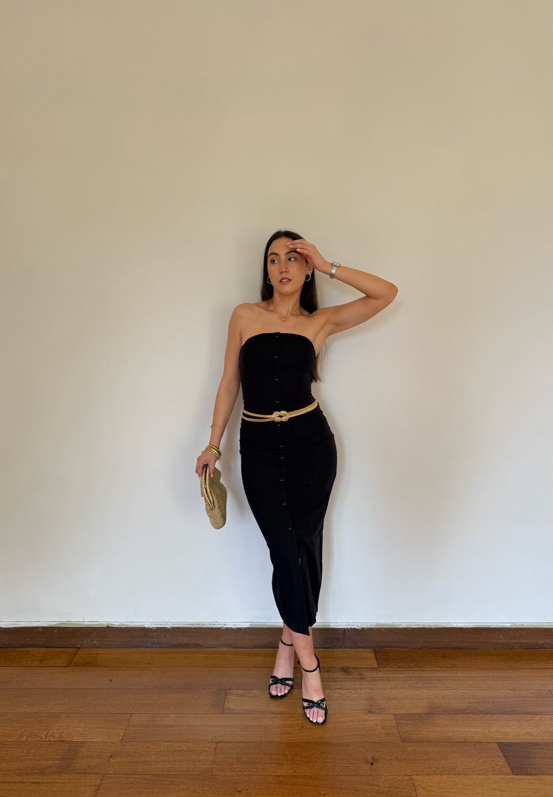 Femme en robe noire bustier avec ceinture beige et pochette, debout sur un sol en bois contre un mur clair uni, posant avec la main sur le front.