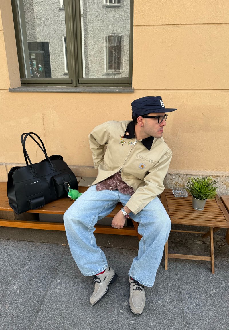 Jeune homme portant une veste beige et une casquette bleue assis sur un banc en bois à côté d'un sac noir et d'une petite table avec une plante.