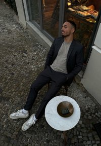 Ciemny garnitur w paski, jasnoszara koszulka, białe trampki z teksturowaną podeszwą, okrągły marmurowy stół z mosiężną misą i małymi książkami. Sceneria na zewnątrz.