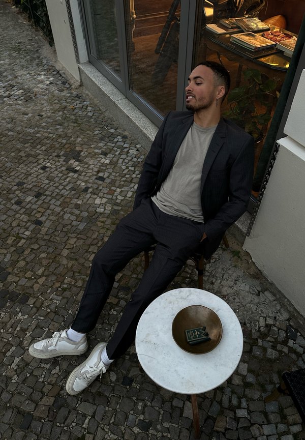 Donker gestreepte kostuum, lichtgrijs T-shirt, witte sneakers met textuurrand, ronde marmeren tafel met een koperen schaal en kleine boeken. Buitenomgeving.