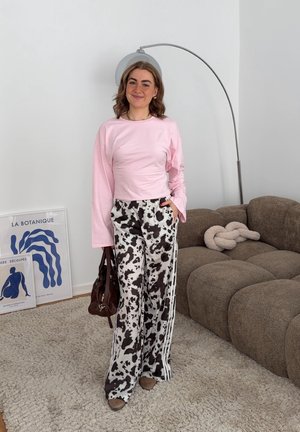 Mujer joven de pie en el interior, vistiendo una camiseta de manga larga rosa y pantalones con estampado en blanco y negro, sosteniendo un bolso marrón junto a un sofá marrón.