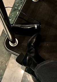 Botas de tornozelo em couro preto com um salto robusto, detalhe de fecho dourado, acompanhadas de uma pequena bolsa preta com fecho. Superfícies do chão texturizadas são visíveis.