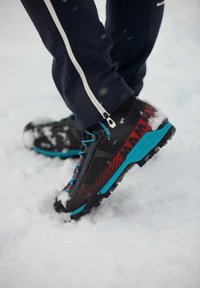 Chaussures de randonnée noires et rouges avec des accents bleus, dotées d'une tige texturée, d'une semelle robuste et de neige sur l'avant, posées sur une surface enneigée.