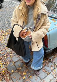 Beige Fleece-Mantel mit braunen Besätzen, grauer Rippenpullover, Jeans mit weitem Bein, braune Stiefeletten und eine schwarze Tragetasche mit Zeitschriften.