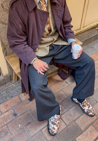 Μωβ παλτό, καρό πουκάμισο, πορτοκαλί ρίγες γραβάτα, γκρι παντελόνι φαρδιάς γραμμής και παπούτσια με σχέδια σε καφέ και λευκό. Κρατάει ένα κύπελλο καφέ.