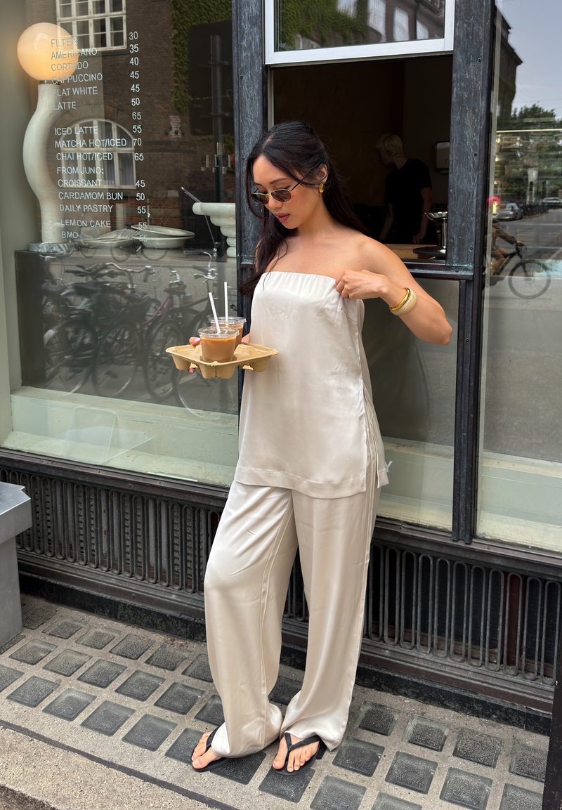 Femme en tenue satinée crème et lunettes de soleil tenant un plateau avec des cafés glacés, debout devant une fenêtre de café avec un menu visible.