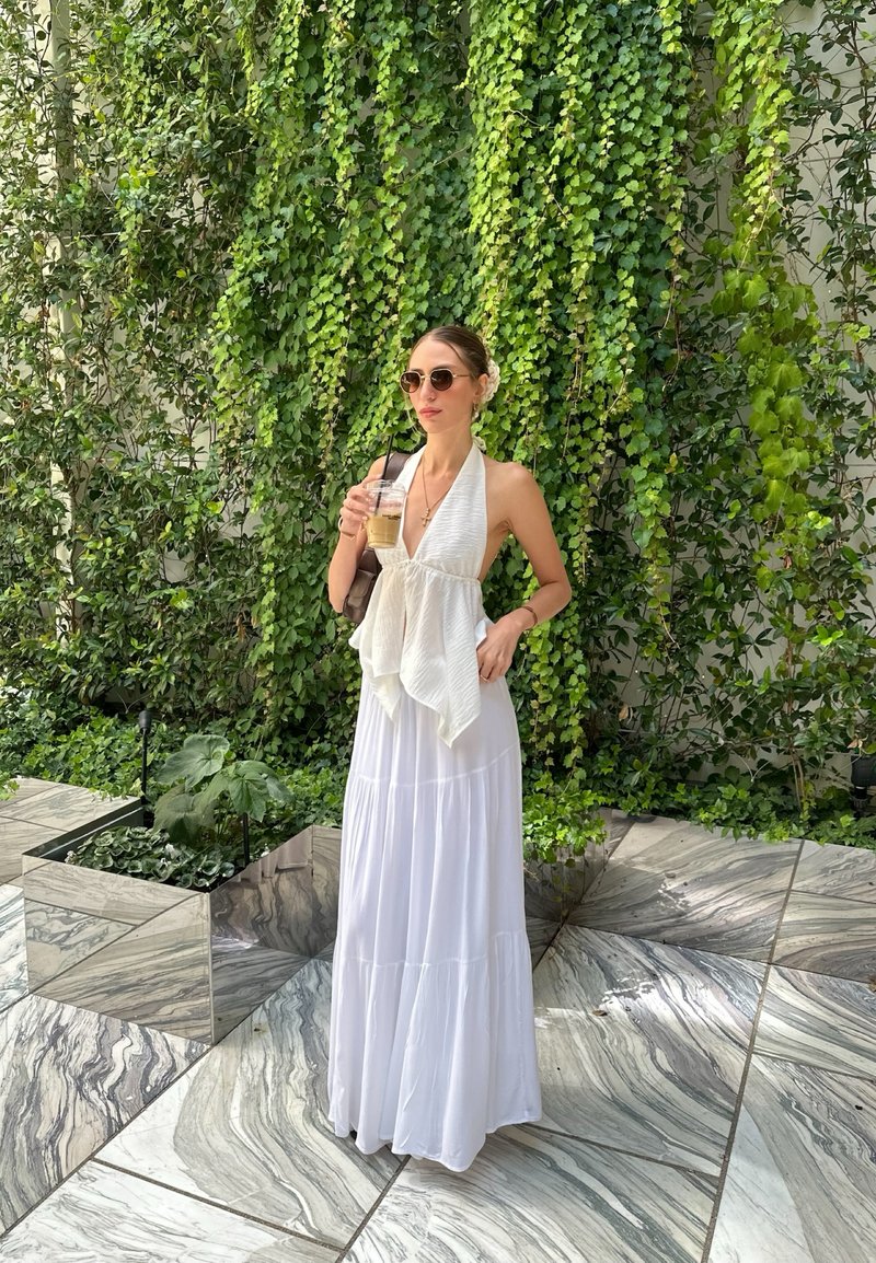 Femme en haut blanc à licou et jupe longue tient un café glacé, debout sur un sol en marbre avec des plantes vertes à feuilles sur le mur derrière elle.
