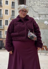 Άτομο που φοράει εκρού μπερέ, μεγάλα κόκκινα γυαλιά, μωβ χνουδωτό μπουφάν, ταιριαστό φόρεμα και μικρή καρό τσάντα χιαστί, στέκεται έξω.