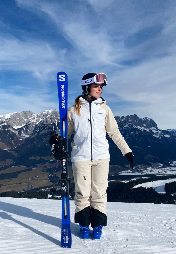 Skiredskabet inkluderer blå Salomon-ski og lysblå skistøvler. Outfitten har en hvid og tan vandtæt skijakke og tan skibukser.