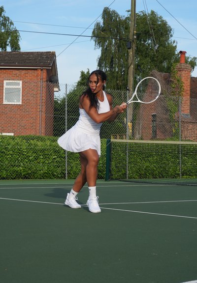 Jugadora de tenis con un vestido blanco sin mangas y zapatos blancos, golpeando una raqueta en una cancha verde con una valla y edificios al fondo.