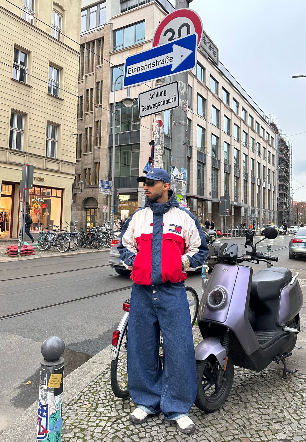 Homme portant une veste rouge, blanche et bleue et un jean large, debout sur un trottoir en ville près d'un scooter violet et de deux panneaux de signalisation en allemand.