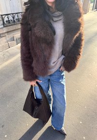 Femme portant un manteau en fourrure brun foncé, un pull beige, un jean bleu, des chaussures noires à bout pointu, tenant un grand sac à main noir tressé dans une rue de la ville.