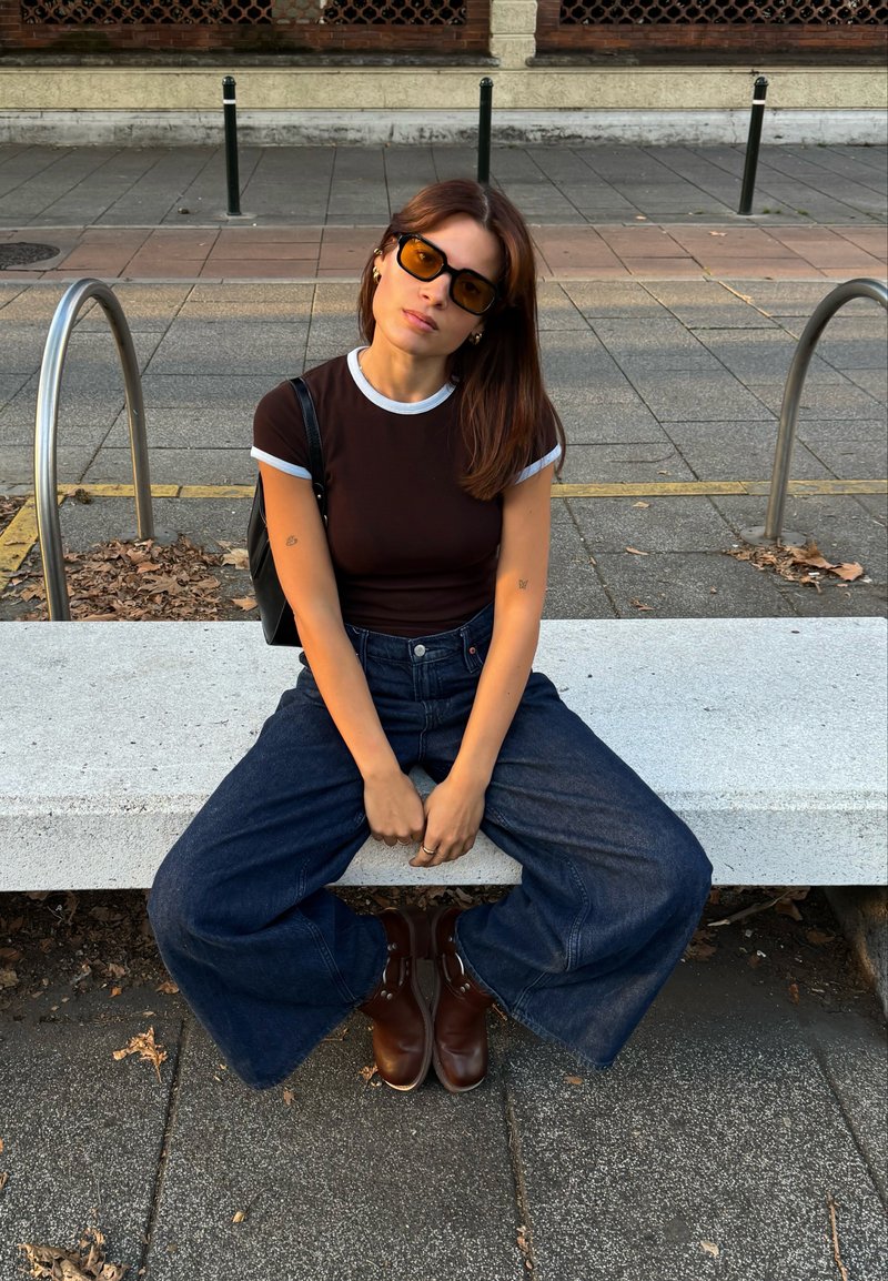 Jovem mulher a usar óculos de sol âmbar, t-shirt preto e branco, jeans largos azuis e botas castanhas senta-se num banco de pedra numa zona urbana.