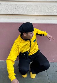 Sudadera amarilla con cremallera y logo, pantalones negros y zapatos amarillos. La persona lleva un boina negra y una pequeña bolsa negra, agachada contra una pared.