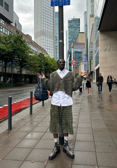 Suéter de ganchillo de color oliva y de corte oversize; camisa blanca con botones; pantalones cortos de cuadros hasta la rodilla; botas de plataforma negras; bolso oscuro; entorno urbano.