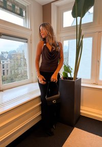 Top maro cu guler cowl și accesorii argintii, pantaloni negri mulați și o geantă mică neagră cu accente aurii, în fața unei ferestre cu plante.