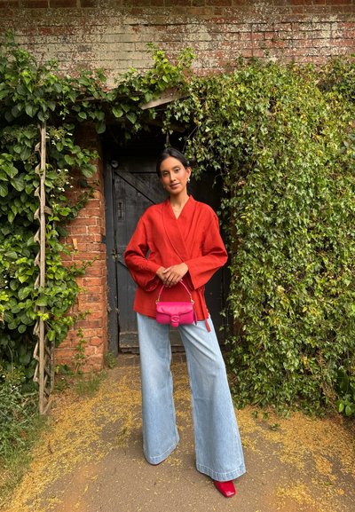 Top envolvente de lino rojo, vaqueros de mezclilla claros de pierna ancha, bolso rosa intenso, tacones rojos brillantes, fondo de follaje verde, pared rústica de ladrillo.