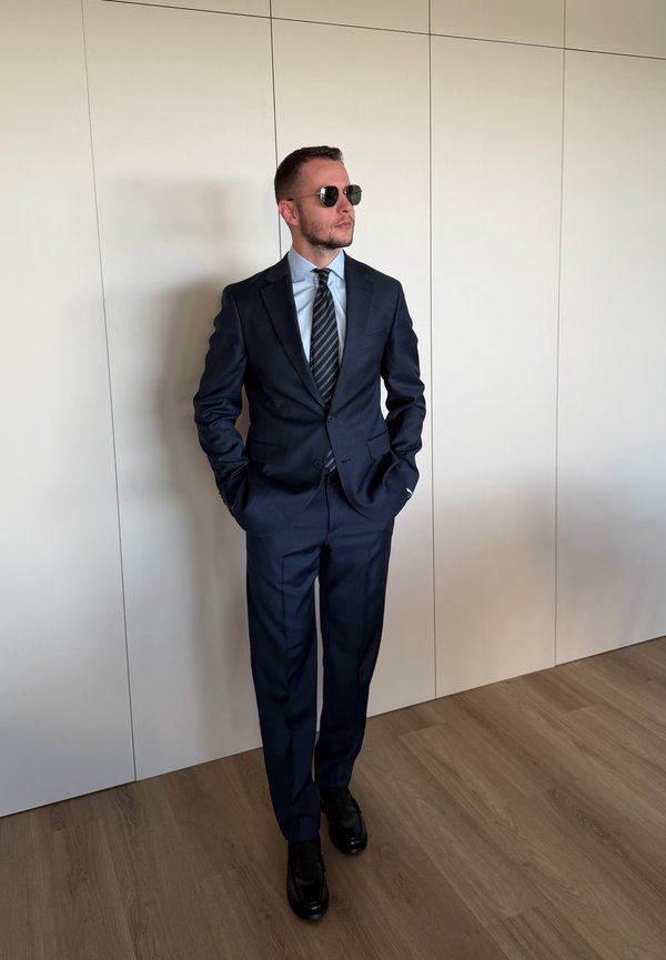 Hombre con traje azul oscuro, camisa azul clara, corbata a rayas, zapatos negros y gafas de sol, de pie en el interior con las manos en los bolsillos contra una pared blanca.
