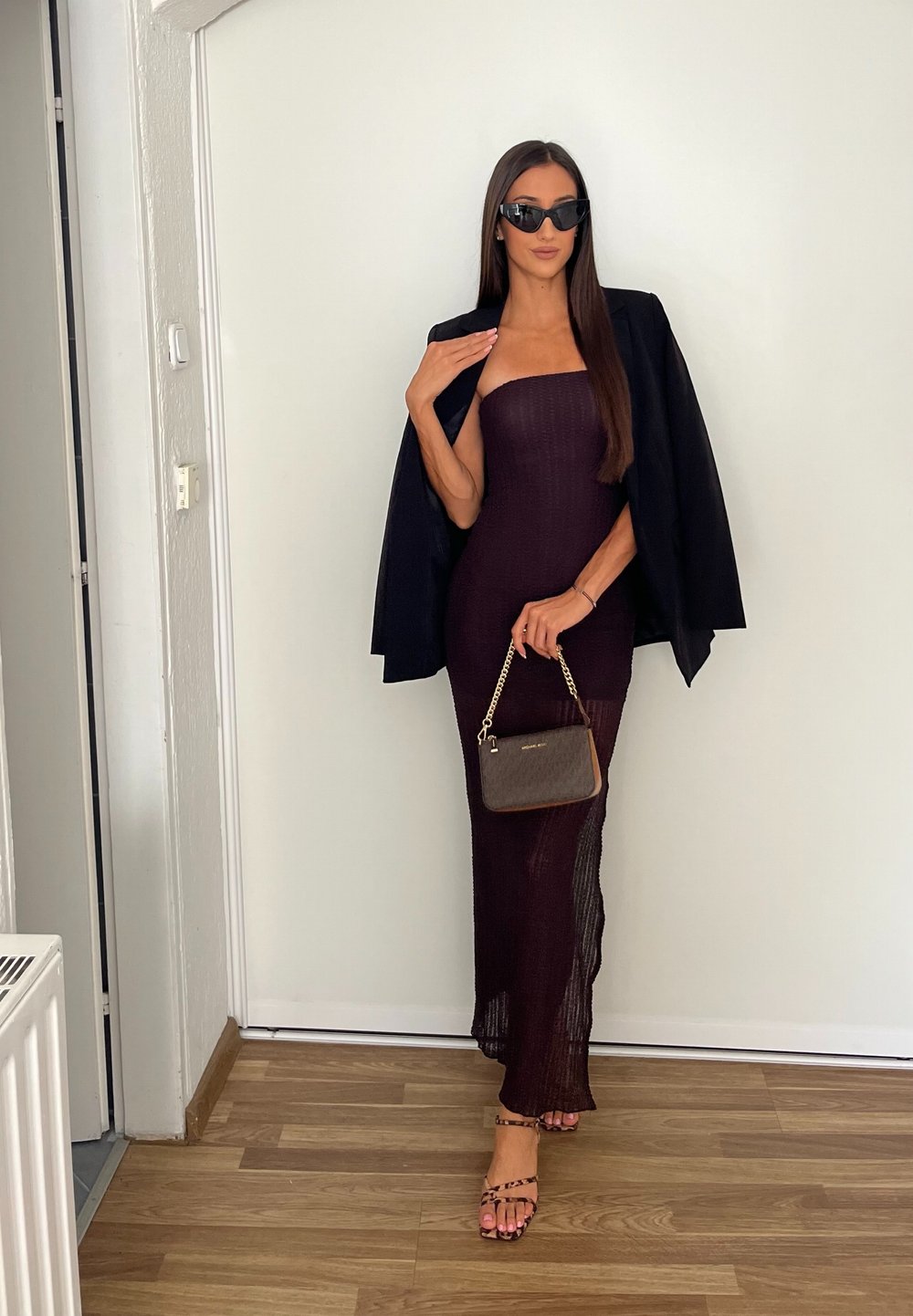 Rochie bordo fără bretele, ajustată, cu țesătură texturată, asortată cu un sacou negru și o geantă mică maro cu lanț de aur. Ochelari de soare negri.