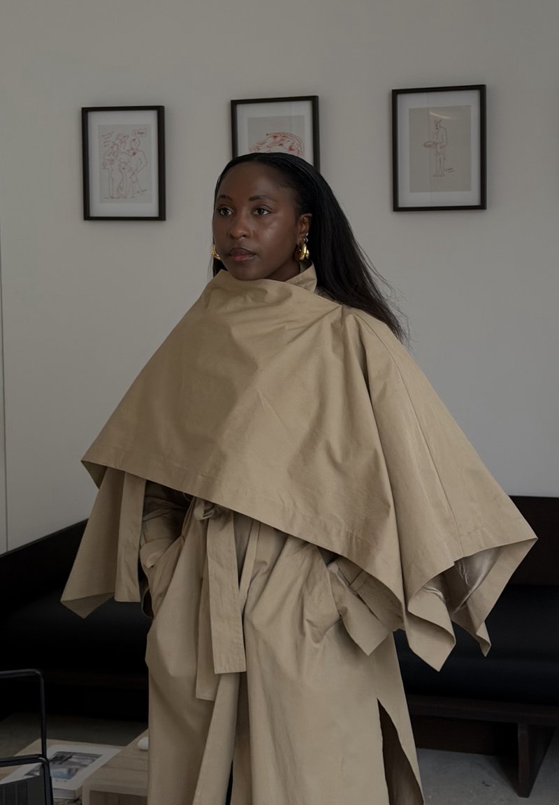 Femme noire portant un manteau cape beige à couches, debout à l'intérieur avec une œuvre d'art minimaliste encadrée sur le mur derrière elle.