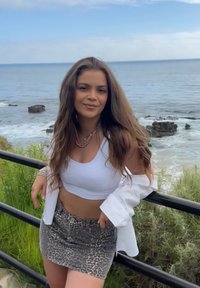 White cropped tank top, grey leopard-print denim skirt, layered necklaces, long wavy brown hair, standing by a railing with ocean in background.