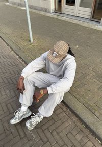 Ljusgrå bomullsoverall med ribbade muddar, brun rutmönstrad keps, silverfärgade löparskor och ett guldur. Asfalterad gata i bakgrunden.