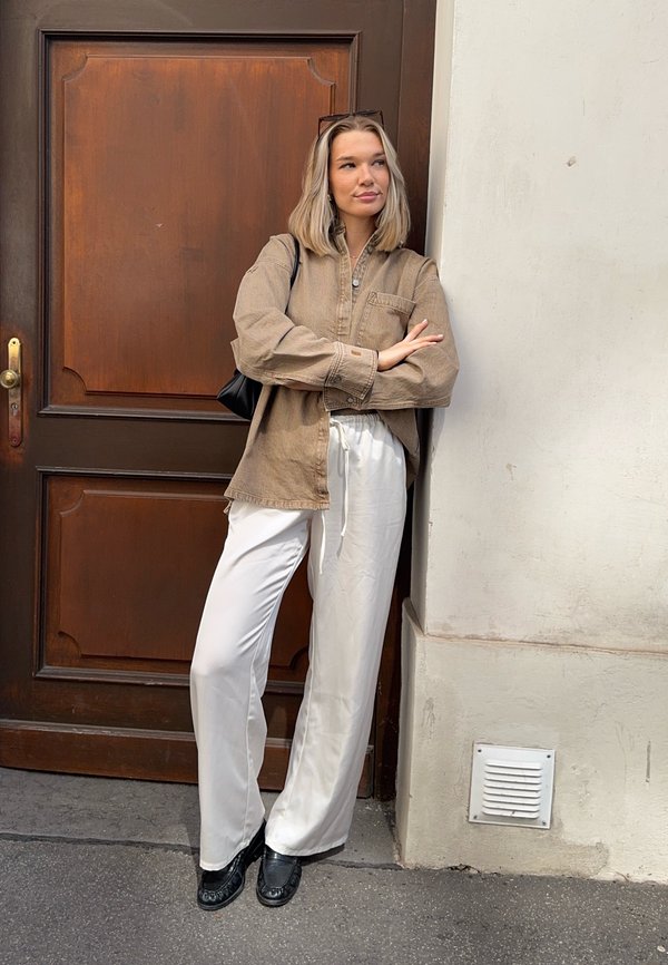 Una mujer con una camisa beige y pantalones blancos se apoya en una pared junto a una puerta de madera, con los brazos cruzados y un bolso negro al hombro.