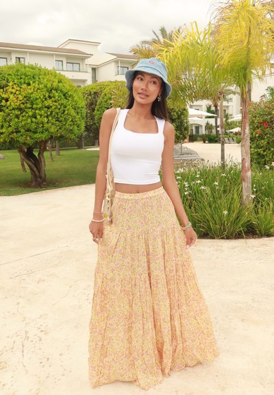 Top corto blanco y una falda maxi floral amarilla con acentos rosas, combinados con un sombrero de cubo azul claro y una bolsa de mimbre.