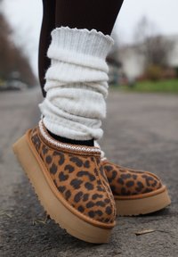 Nohy v leopardích nazúvacích topánkach s hrubými podrážkami a bielymi nariasenými návlekmi na nohy na dláždenej vonkajšej ceste.