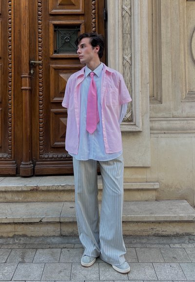 Camisa rosa claro de manga corta sobre una camisa azul de botones, combinada con pantalones de pierna ancha a rayas. El modelo lleva una corbata rosa y zapatos de slip-on. Fondo de puerta de madera oscura.