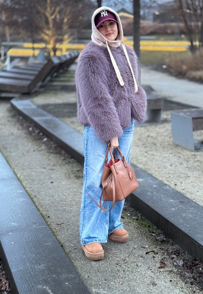 Mujer de pie al aire libre con chaqueta de piel sintética morada, jeans azules, botas color beige, gorro de punto beige con cordones y gorra morada, sosteniendo un bolso marrón.